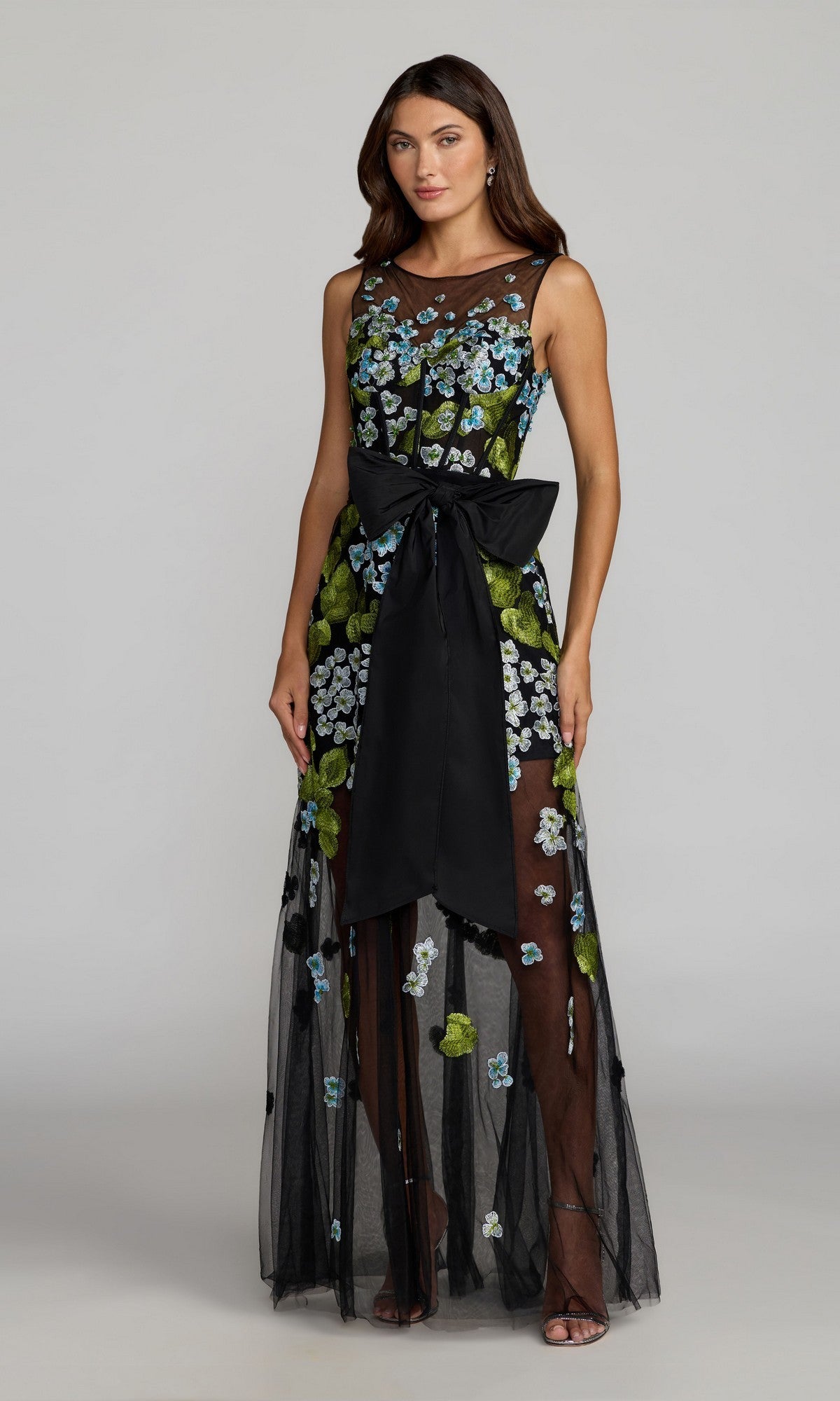 A woman stands against a plain background in the Mac Duggal 21058 long formal dress, featuring a sleeveless blue multi design with floral embroidery and a statement waist bow.